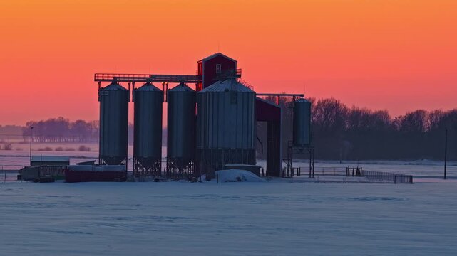 Aerial view of a grain silo standing in a snow-covered field during a vibrant sunset in Europe.