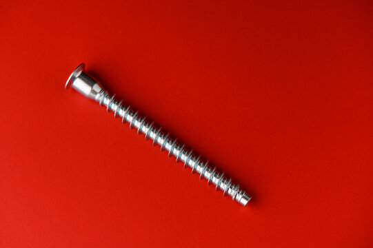 This striking metal screw rests flat against a lively red background, showcasing its intricate thread and glossy finish. The contrast between the shiny silver and bold red creates a visually appealing