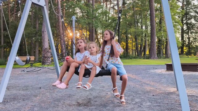 Three girls enjoy their time at the swings in a park. They smile and chat while two girls push the swing for the youngest girl