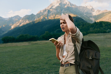 Obraz premium A young woman with a backpack stands in a mountain field holding a smartphone and looks confused while touching her head. Concept of outdoor lifestyle and navigation challenges.
