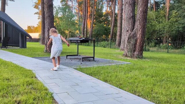 A child walks carefully on a stone path in a park surrounded by tall trees. A grill is visible in the background as the sun sets