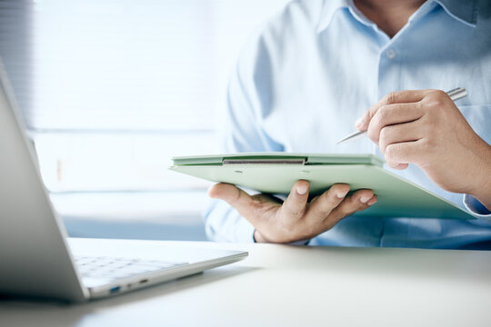 Business professional using clipboard and pen reviewing documents at office desk beside laptop computer, close-up hands managing digital paperwork, workflow planning, corporate productivity
