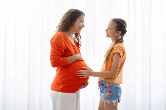 Pregnant mother and young daughter standing by bright window, smiling and bonding, female child touching baby bump while sharing excitement, affection, and anticipation for the new baby