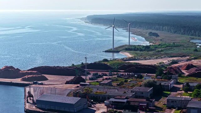 Aerial view of industrial port with wind turbines, storage piles, and coastal forest, Europe.