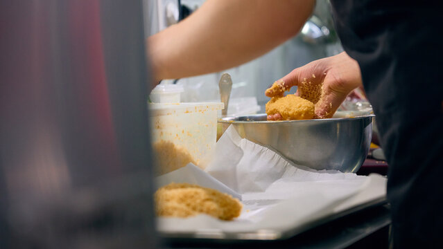 Food worker portioning breaded mixture into metal bowl in professional kitchen. Concept of manual food processing, portion control, restaurant prep station workflow.