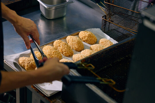 Kitchen worker loading breaded portions into fryer basket during food production. Concept of high volume cooking, restaurant back of house, standardized meal production.