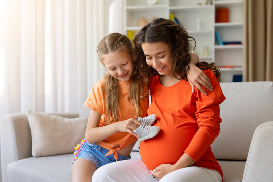 Excited daughter holding tiny baby shoes beside her pregnant mothers belly as they smile together, preparing with love and anticipation for a new family member in their bright home