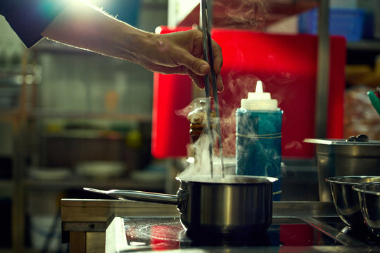 Kitchen worker adding ingredient to steaming saucepan during food preparation. Concept of recipe execution, ingredient control, commercial kitchen operations.