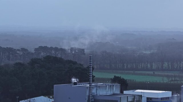 Aerial view of an industrial building with a smoking chimney surrounded by trees and fields in Europe.