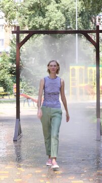 Smiling woman enjoying refreshing cooling mist under outdoor water spray on hot summer day in city park. Joyful moment, raised hands, relief from heat, healthy active lifestyle, urban recreation, copy