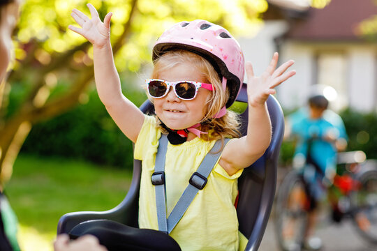 Portrait of little toddler girl with security helmet on the head sitting in bike seat of parents. Boy on bicycle on background. Safe and child protection concept. Family and weekend activity trip.