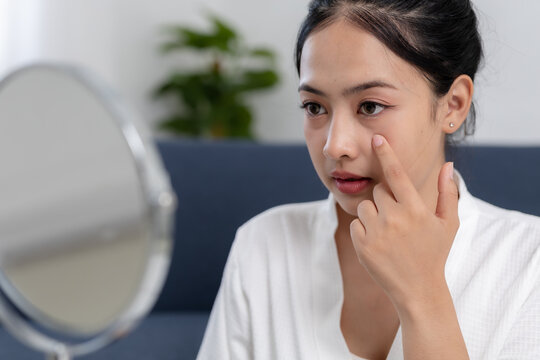 Concerned young asian woman looking at reflection in mirror touching face under eye checking skin problem with sad worried expression during daily morning beauty care routine at home
