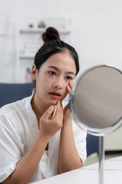 Concerned young asian woman looking into round mirror checking her face for skin blemish during daily personal care routine feeling worried about dermatological issues