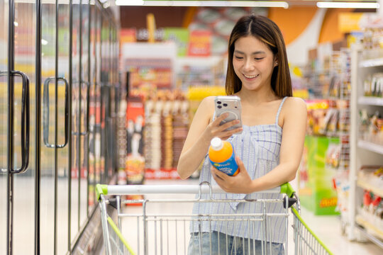 Asian woman use smartphone to scan a product QR code to access information while push shopping cart down an aisle to buy food and beverage in supermarket. Beautiful customer select products in fridge.