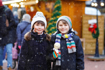 Obraz premium Little cute kid girl and boy eating white chocolate-covered strawberries and apple skewers at German Christmas market. Happy siblings. Real life, real people, snowy festive joy