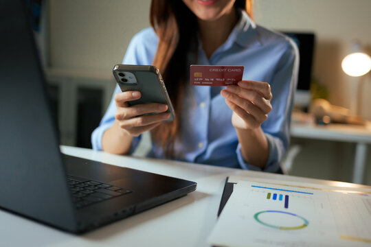 Woman holding a credit card and smartphone for online transactions, shopping, or secure payment, with a laptop and business documents on a desk