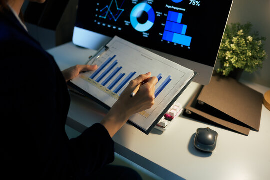 Person reviewing business reports and graphs on a clipboard while comparing information with digital data on a computer screen
