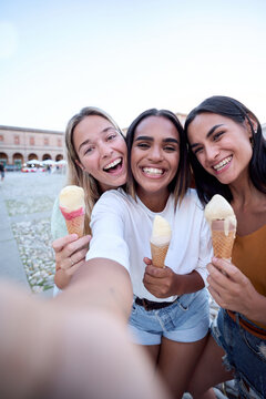 Vertical. Three cheerful female friends are enjoying a summer day, taking a selfie while laughing and eating ice cream cones in a picturesque town square