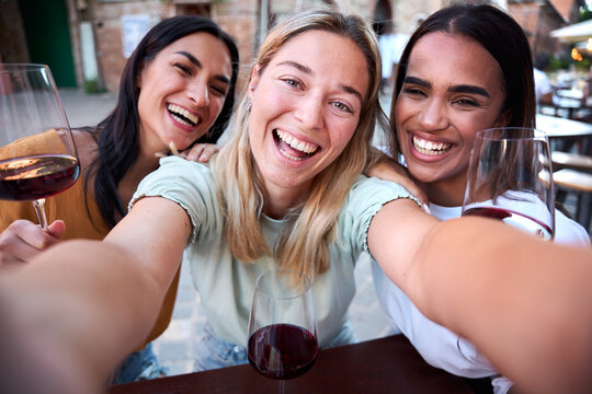 Three cheerful multiracial female friends are smiling and taking a selfie while drinking glasses of red wine at a restaurant in a charming european town
