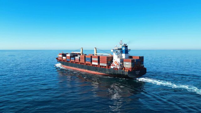 Large container cargo ship sailing on calm blue ocean under clear sky, leaving wake trail