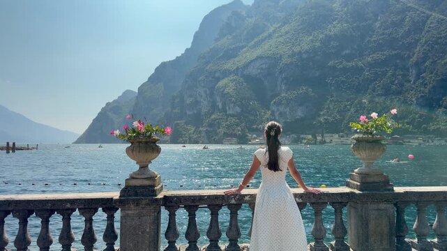 Woman Admiring Lake Garda from Scenic Promenade in Riva del Garda, Rear View of Elegant Female Traveler in White Dress with Turquoise Water and Dramatic Alpine Mountains in Northern Italy