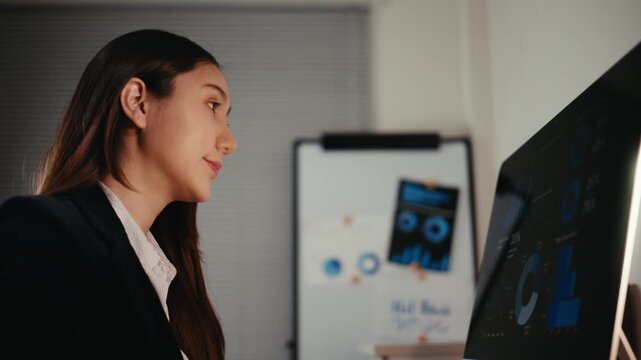 Young Asian business analyst typing and looking at computer screen with charts and graphs, working hard late in a modern office