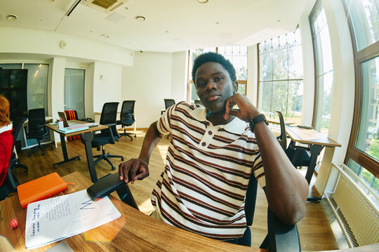Portrait of young adult Black man sitting at office desk looking into camera. Image represents corporate diversity and modern workplace culture, suitable for HR recruitment campaigns