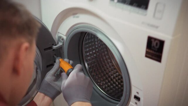 A technician in uniform and gloves removes a screw from a washing machine drum under neutral bathroom lighting. Concept for home appliance maintenance and diagnostics