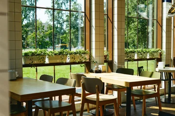 Fotobehang Horeca Empty wooden tables and chairs in modern cafe with large windows showing green park outside, representing hospitality industry and dining concepts, perfect for restaurant marketing  © Seventyfour