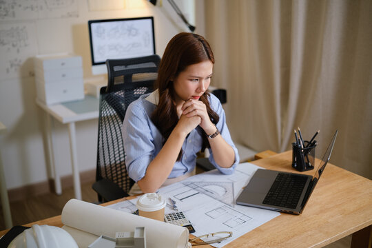 Young female architect working at a desk with laptop, blueprints, calculator, and coffee in a modern home office.