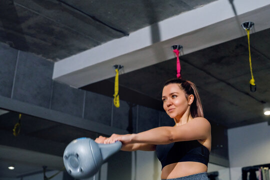 Young woman performing kettlebell swing with focused strength. Female athlete training explosive power in modern gym, fitness workout concept.