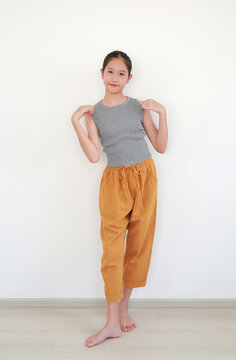 Pretty young Asian girl in ribbed gray top and rolled mustard yellow pants smiles while playfully lifting the hem of her shirt, posing bare feet on a wooden floor on white wall.