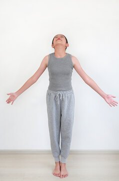 Beautiful young Asian girl in comfortable grey sportswear practicing a graceful yoga pose against white wall in room.