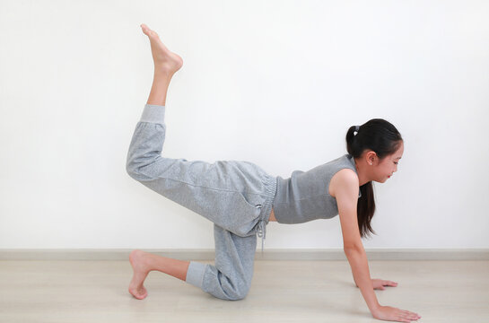 Beautiful young Asian girl in comfortable grey sportswear practicing a graceful yoga pose against white wall in room.