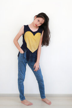 A playful young Asian girl with long flowing hair tilts her head and smiles brightly while posing in navy heart-print top and blue jeans against a white wall.