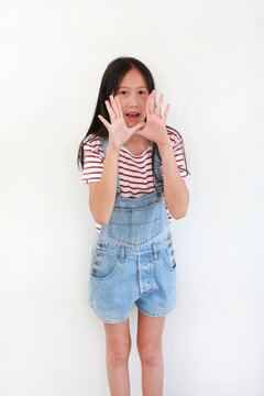 Asian girl in red and white striped t-shirt with denim overalls stands against white background, cupping her hands around her mouth to shout or make an announcement.