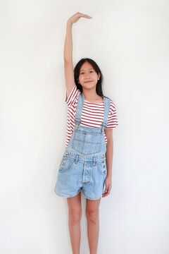 A smiling young Asian girl in denim overalls standing against white studio background, raising one arm straight up above her head to measure her height.