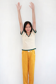 A smiling young Asian girl standing against white studio background and raising both arms straight up in the air.