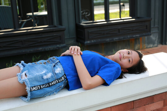 Young Asian girl wearing a blue t-shirt and denim shorts posing lying on bench in the garden.