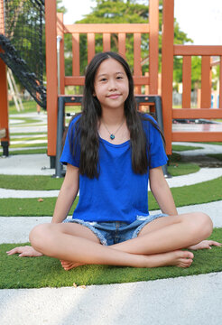 An Asian girl wearing blue t-shirt and denim shorts is sitting cross-legged on artificial grass at a playground and looking at camera with a gentle smile.