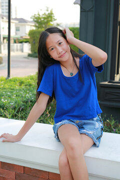 Portrait of Asian girl wearing a blue t-shirt and denim shorts is sitting and smiling on a brick ledge outside during the day.