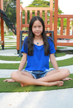 Asian pre-teen girl in casual blue t-shirt and denim shorts, sitting cross-legged and barefoot on the green grass floor of an outdoor playground while smiling gently at camera.