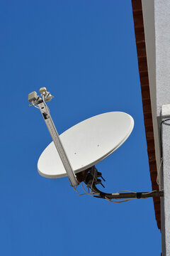 A low angle shot of a white parabolic satellite dish receiver mounted on a white building wall under a clear blue sky.