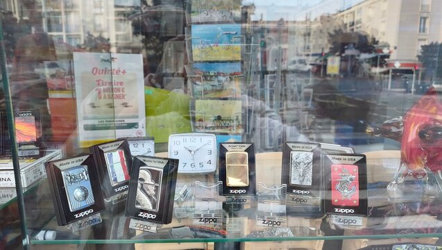 Royan, France - July 15, 2026: Charente-Maritime. Nouvelle-Aquitaine. Display of various Zippo lighters and clock in shop window with colorful postcards and promotional materials visible