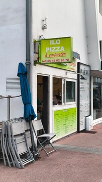 Royan, France - July 15, 2026: Charente-Maritime. Nouvelle-Aquitaine. Ilo Pizza takeout restaurant exterior with green signage, blue umbrella, and stacked chairs in urban setting