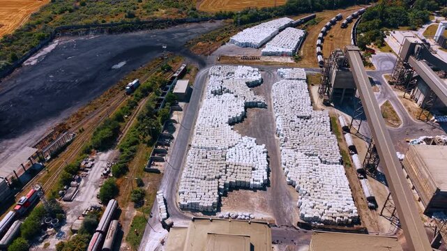 Wrapped materials stored beside industrial rail yard. Large stacks of white packages fill a paved storage area beside freight cars and enclosed conveyors.