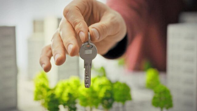 A man's hand holds out shiny keys against a backdrop of 3D models of modern new buildings. Conceptualization of real estate transfers and finalizing a home purchase transaction