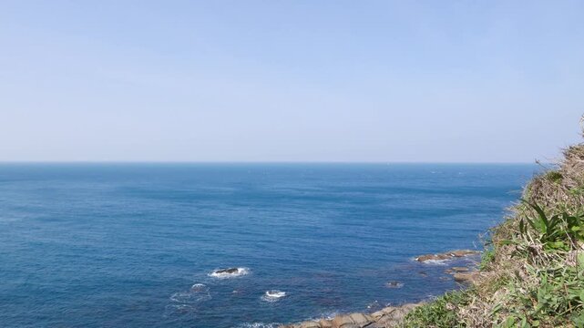 Bird's eye of bluish sea waters and rock shelf from hill peak, grassy area ahead. Camera slowly pan left and tilt down, show outstanding ridge structure, collapsed volcanic creation