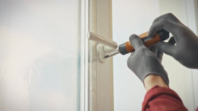 A specialist in gloves adjusts the mechanism of a plastic window with a screwdriver in a home environment. Concept of calling a technician for technical servicing of window hardware
