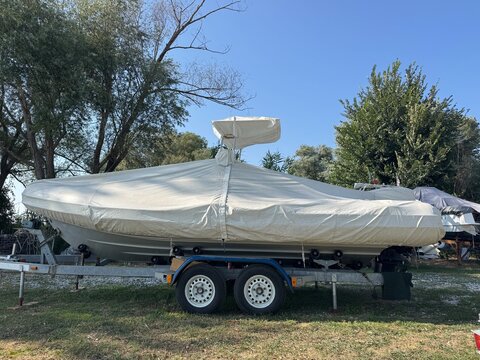 Side view of covered rigid inflatable boat parked on dual axle trailer in grassy outdoor storage area under clear sky. Marine vessel transport concept represents boating maintenance and storage.